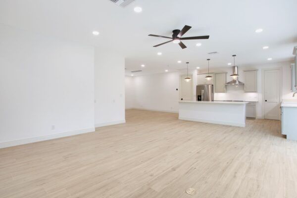 An empty living room with a ceiling fan and a kitchen in the background.