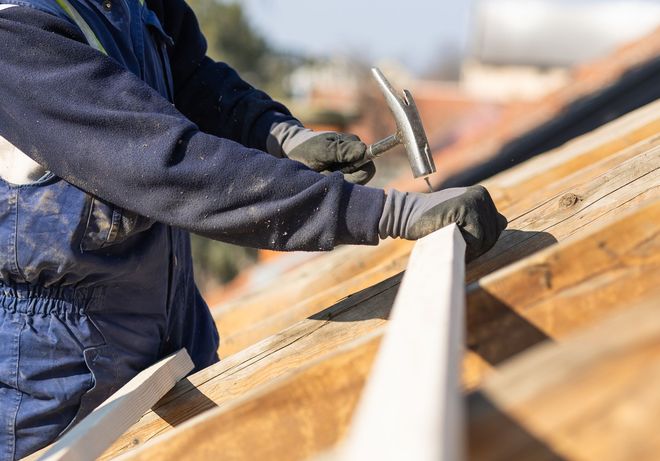 A worker wearing blue overalls and gloves hammers a wooden beam on a roof structure.