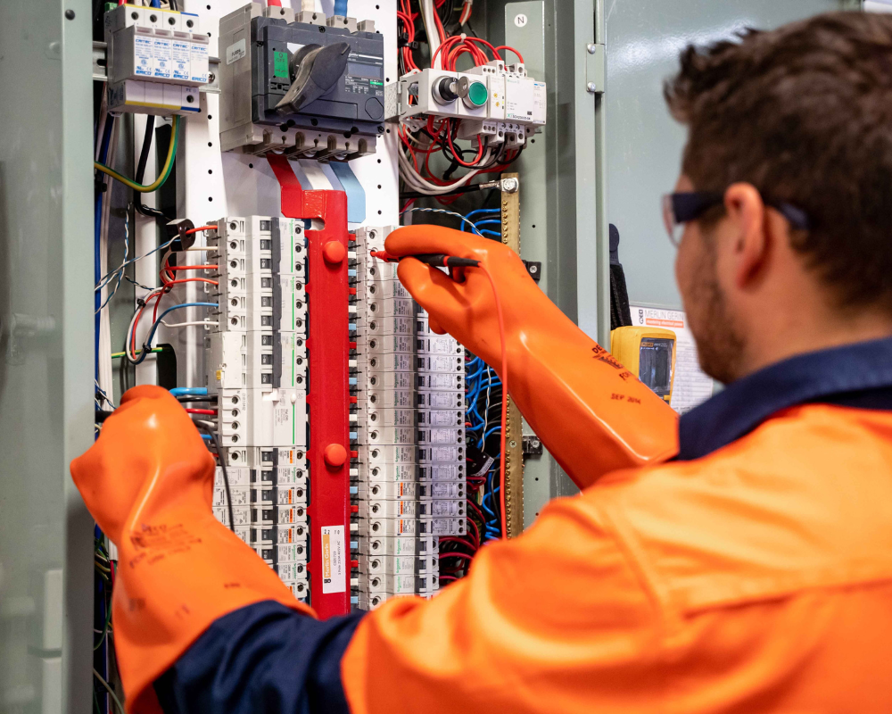 Electrician in orange gloves working on an electrical panel, wires, and equipment.