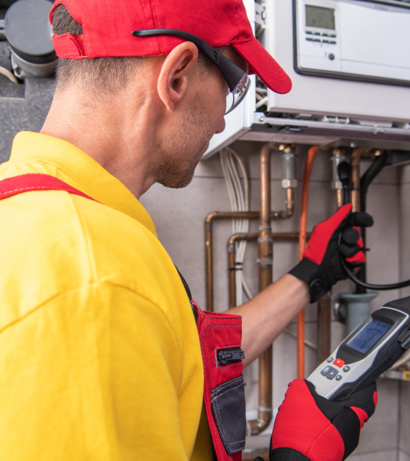 Plumber in yellow uniform and red cap, using a handheld device to inspect a boiler.