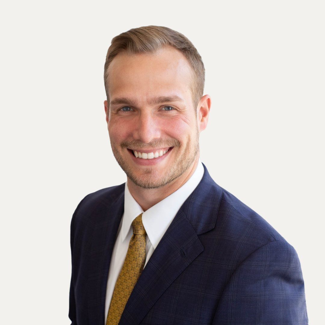 Man in navy suit, white shirt, and gold tie smiling.