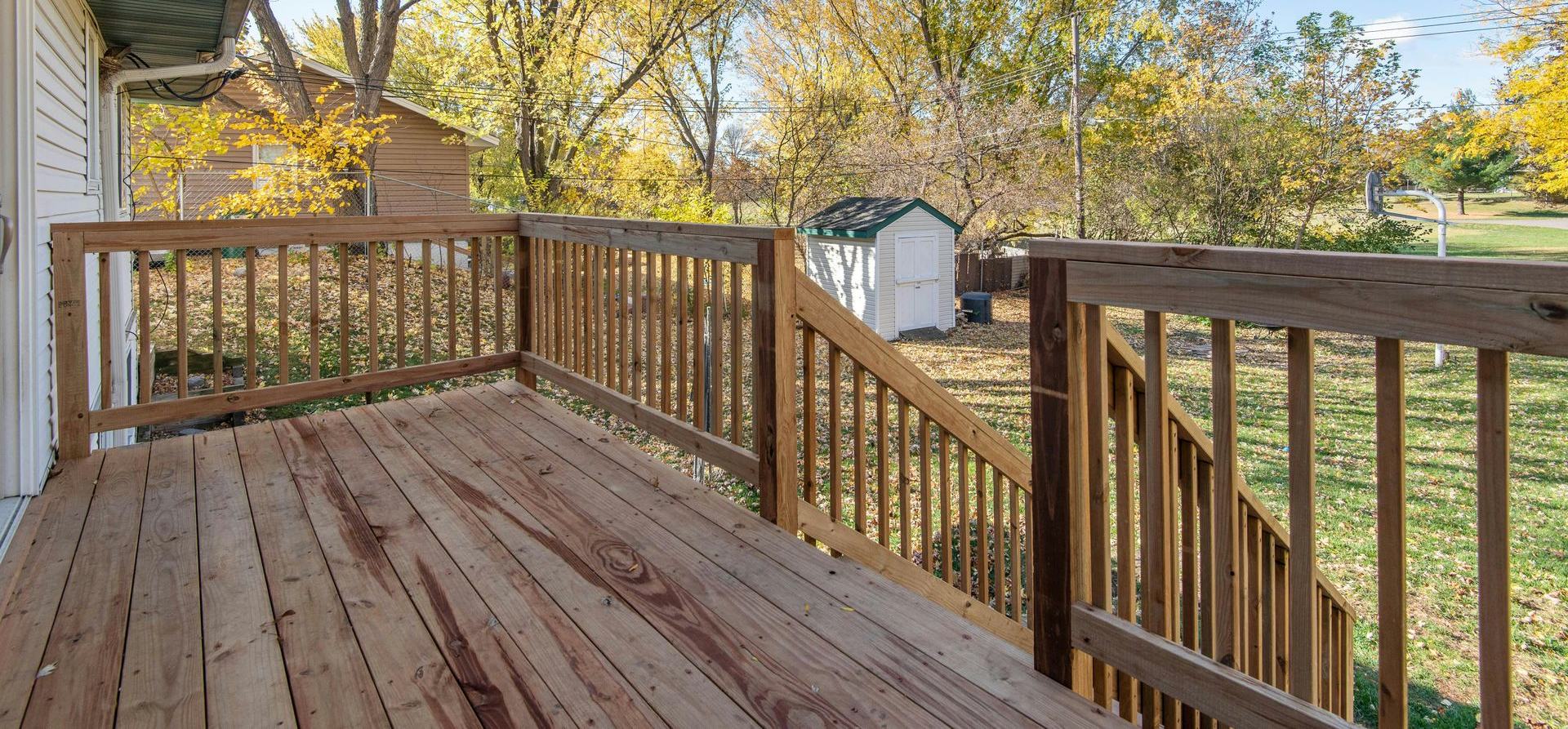 Wooden deck with railing overlooking a yard with trees and a shed on a sunny day.
