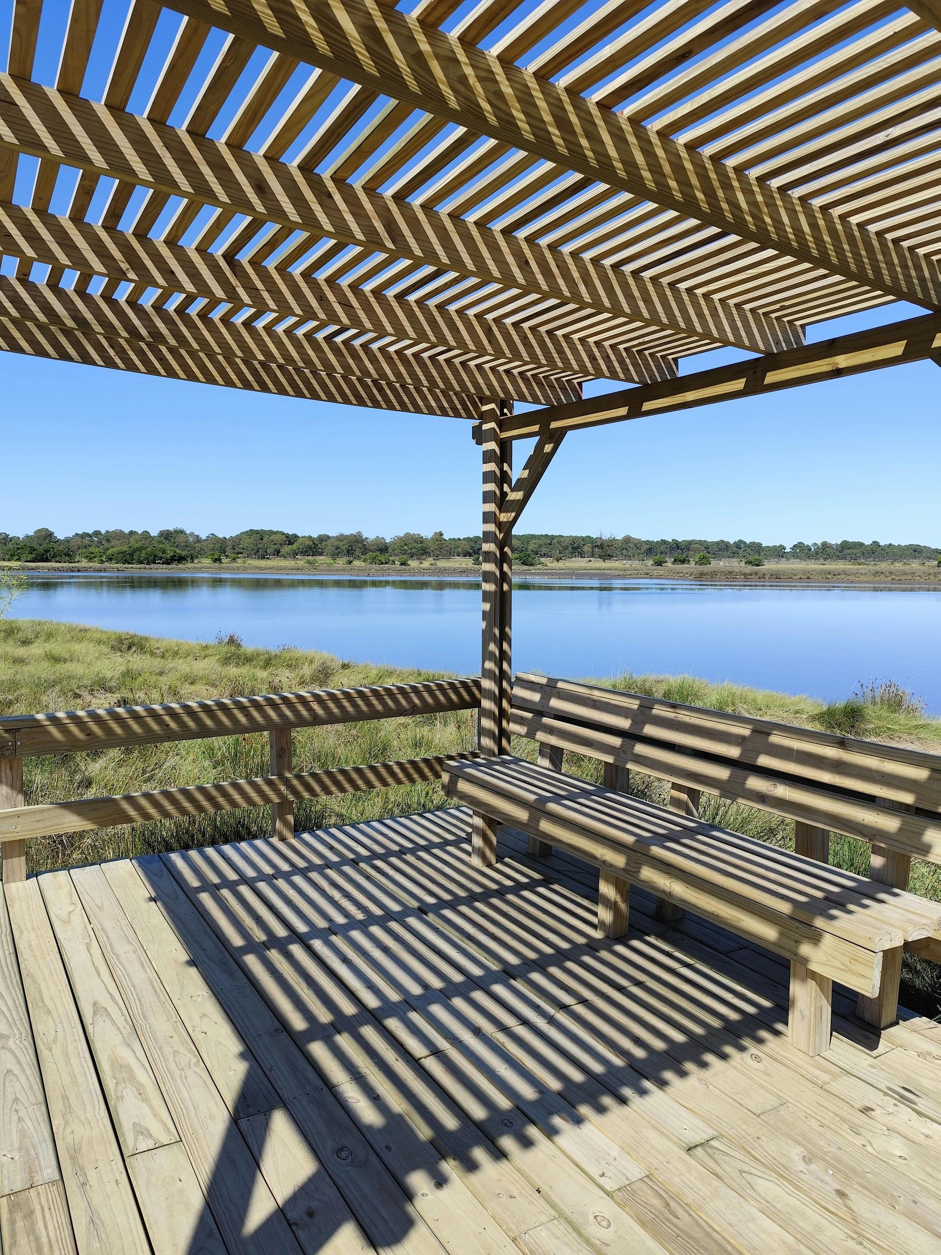 Wooden pergola and bench overlook a lake. Shadows from the pergola create stripes on the deck.