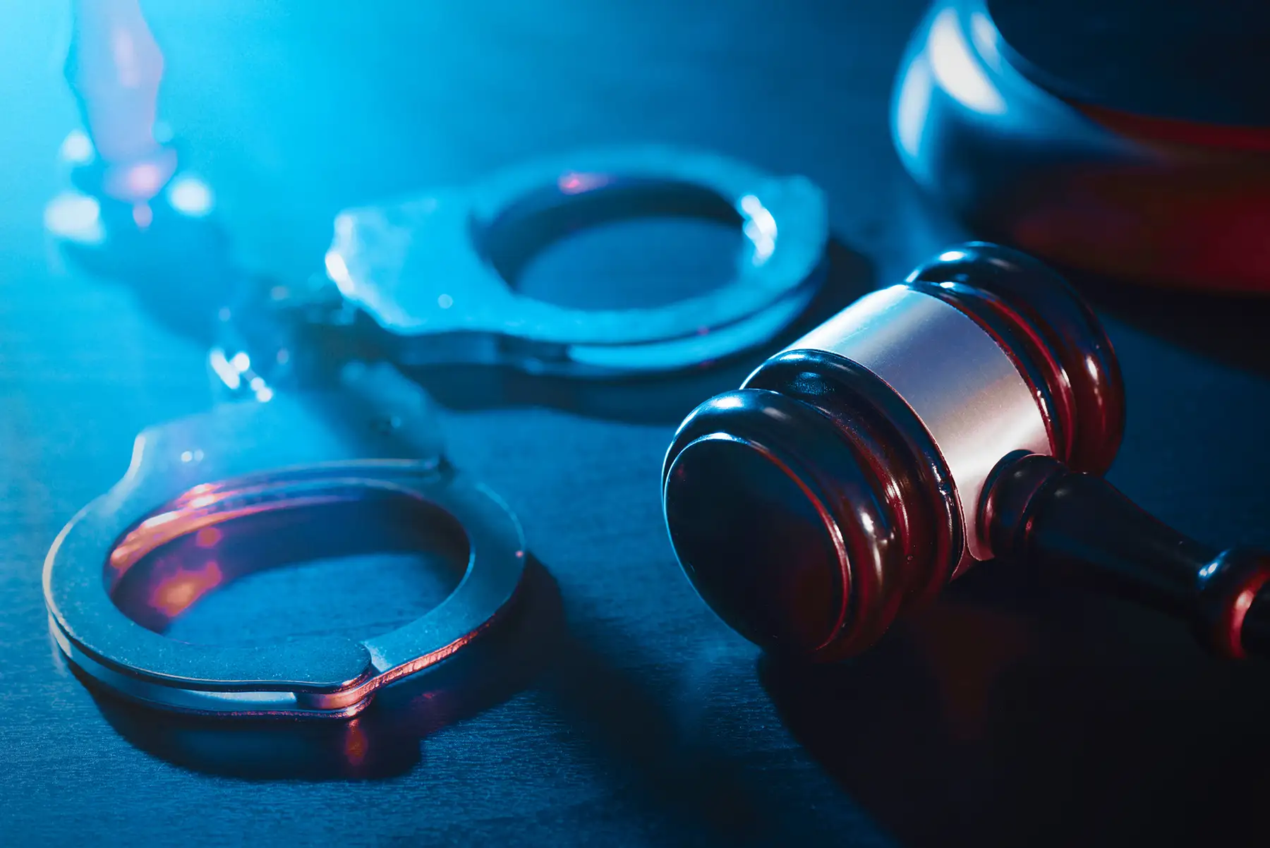 Handcuffs and gavel on a dark surface, lit with blue and red light, suggesting law enforcement or court setting.