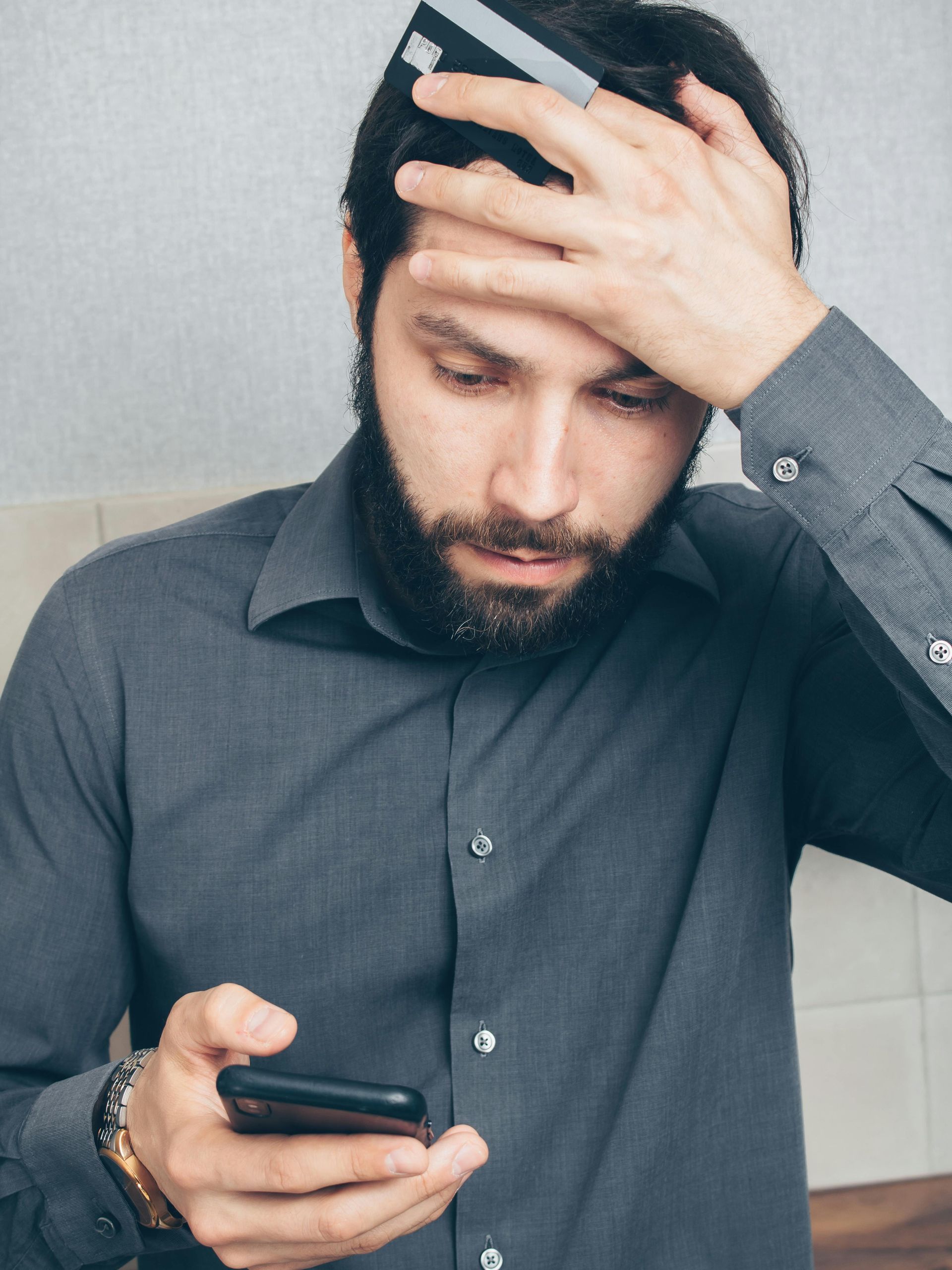 Man in gray shirt holding phone and credit card, looking concerned, hand on head.