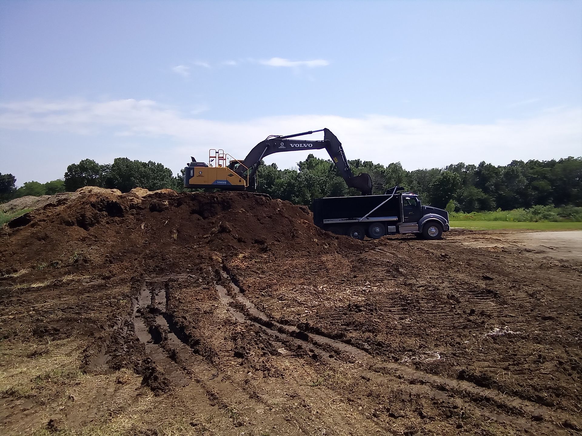 An excavator loading a dump truck with wood chips at an outdoor composting site on a sunny day.