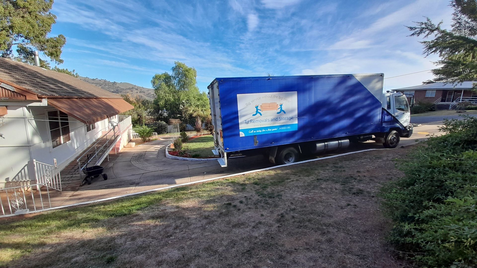 Blue delivery truck parked beside a house in a sunny yard, with a covered load and grassy foreground — TJ Removals and Storage in Wagga Wagga, NSW