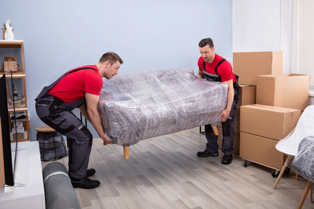 Two Men Are Carrying a Couch Wrapped in Plastic in a Living Room — TJ Removals and Storage in Junee, NSW