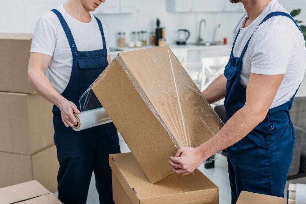 Two Men Are Wrapping a Cardboard Box With Plastic Wrap — TJ Removals and Storage in Wagga Wagga, NSW