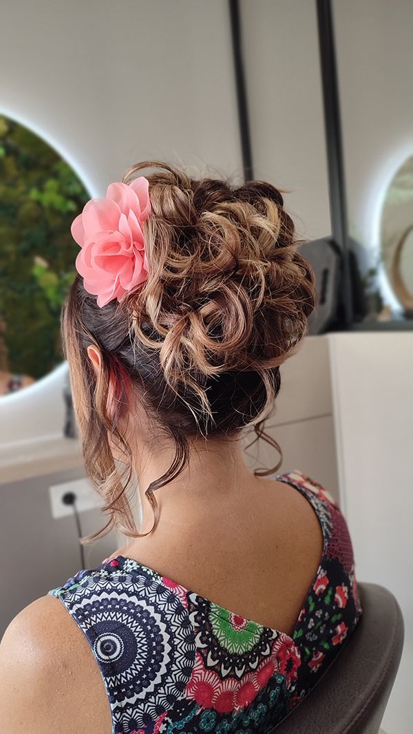 Une femme est assise sur une chaise avec ses cheveux en chignon et une fleur rose dans ses cheveux.