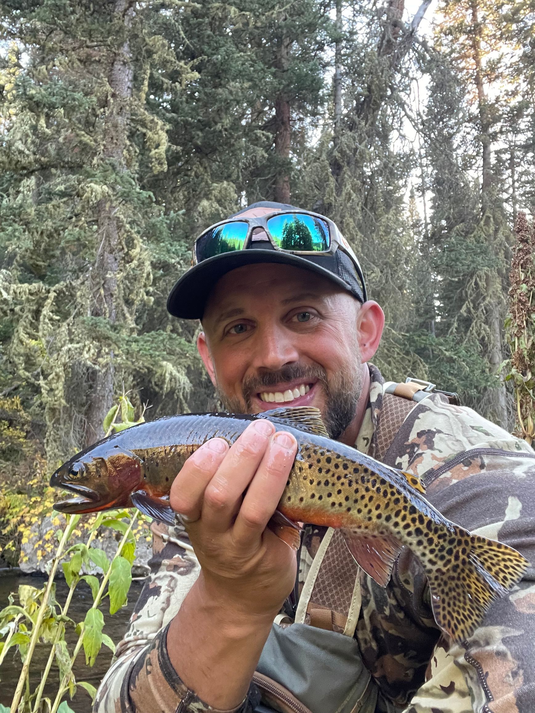 Man in camo holds a trout, smiling near trees.