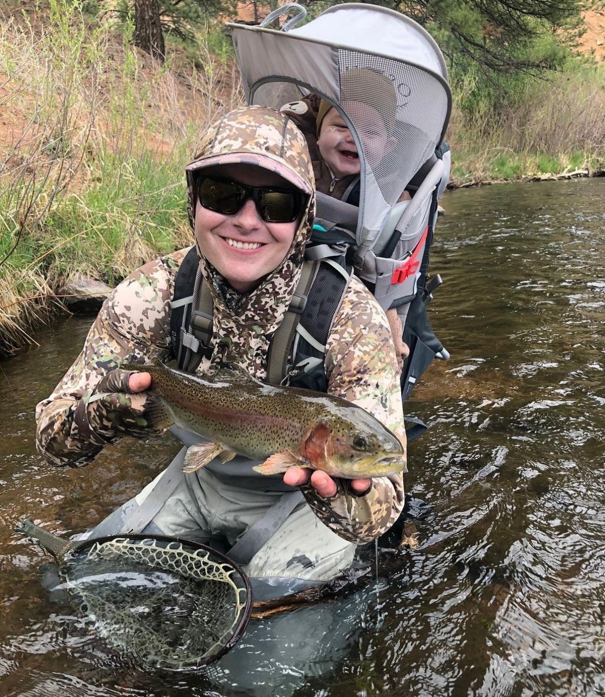 Person holding a fish, wading in a stream with a child in a backpack carrier.