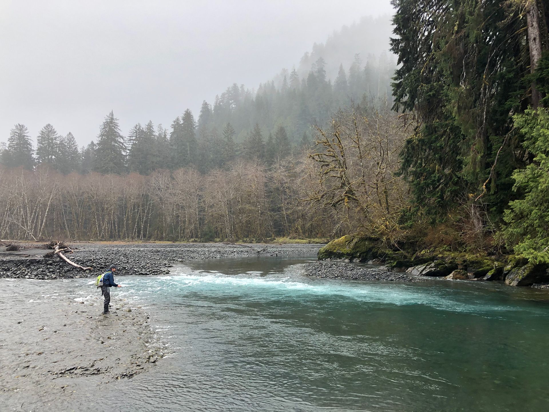 An angler stands on the shore of a misty riverbank. 