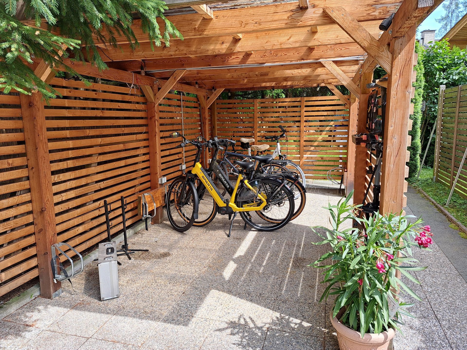 Bikeport: Fahrräder stehen in einem hölzernen Unterstand mit Lattenwänden und Kiesboden. Ein gelbes Fahrrad sticht besonders hervor.