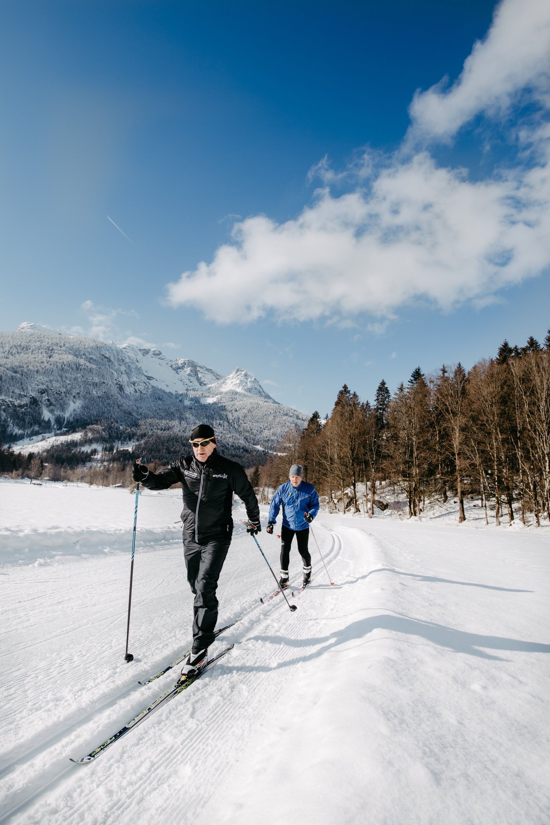 Zwei Personen fahren Langlauf auf einer schneebedeckten Loipe in der Nähe eines Berges und von Bäumen unter blauem Himmel.