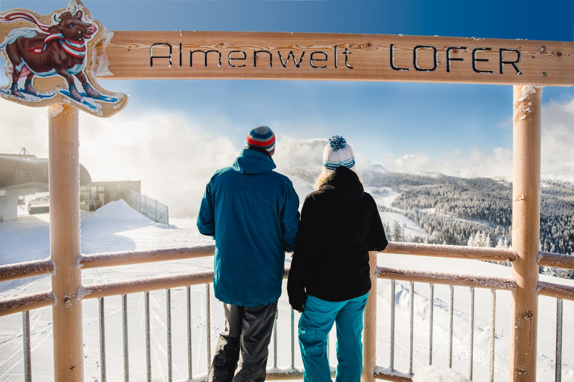 Ein Paar blickt von einem hölzernen Aussichtspunkt auf schneebedeckte Berge, darüber ein Schild mit der Aufschrift „Almenwelt LOFER“.