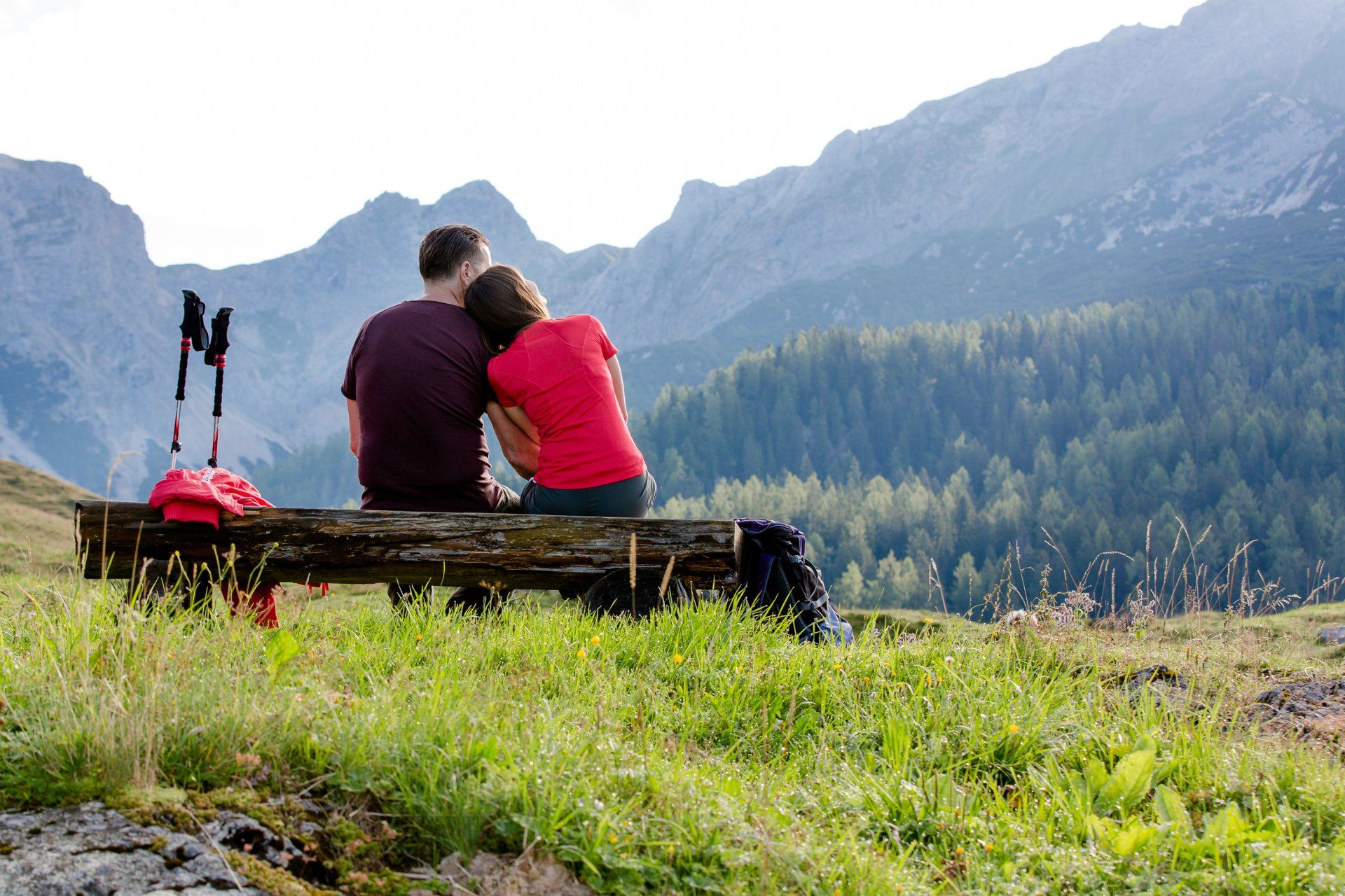 Ein Paar sitzt umarmend auf einer Baumstammbank und blickt über die Berge und einen Wald.