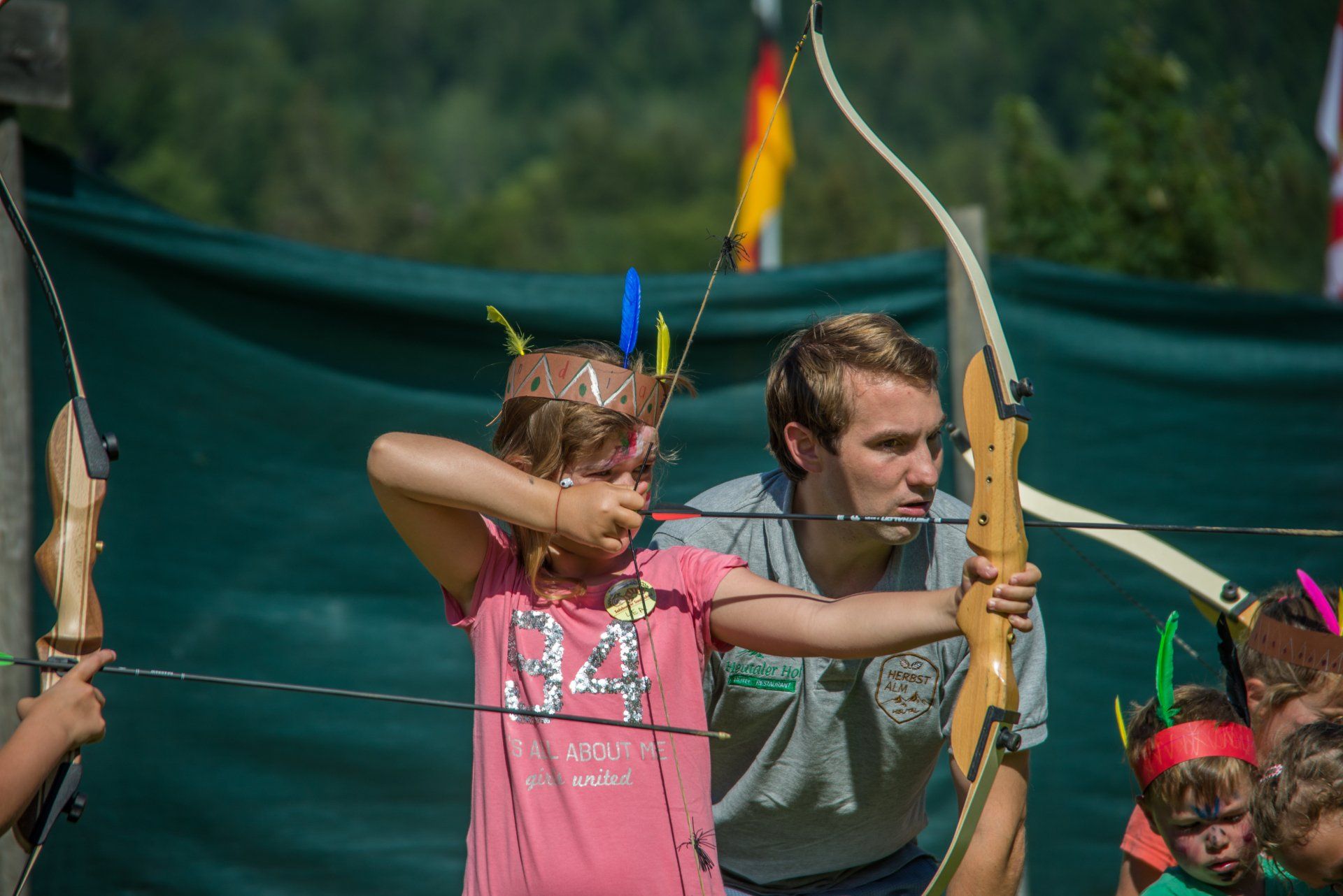 Mädchen mit Federstirnband, das von einem Mann im Bogenschießen unterrichtet wird; beide zielen mit ihren Bögen. Im Freien, grüner Hintergrund.