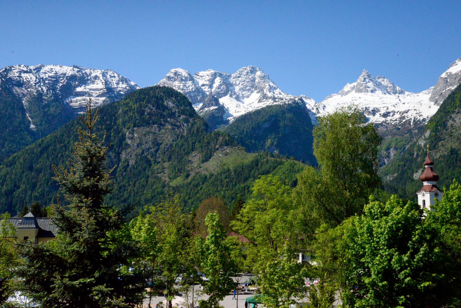 An einem sonnigen Tag erheben sich schneebedeckte Berge hinter grünen Wäldern und einem Dorf mit Kirche in den österreichischen Alpen.