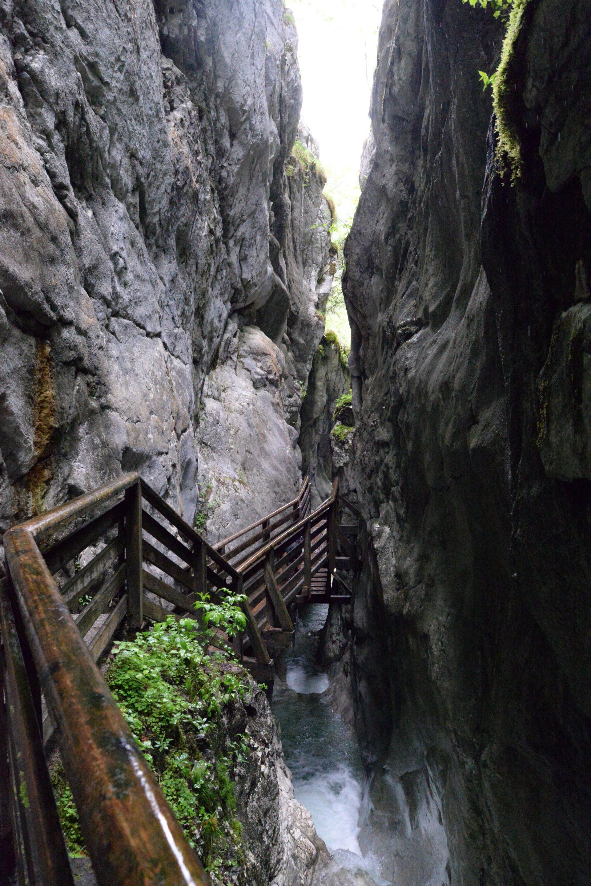Eine Holztreppe führt hinab in eine enge Schlucht mit reißendem Wasser. Steile, graue Felswände säumen den Weg.