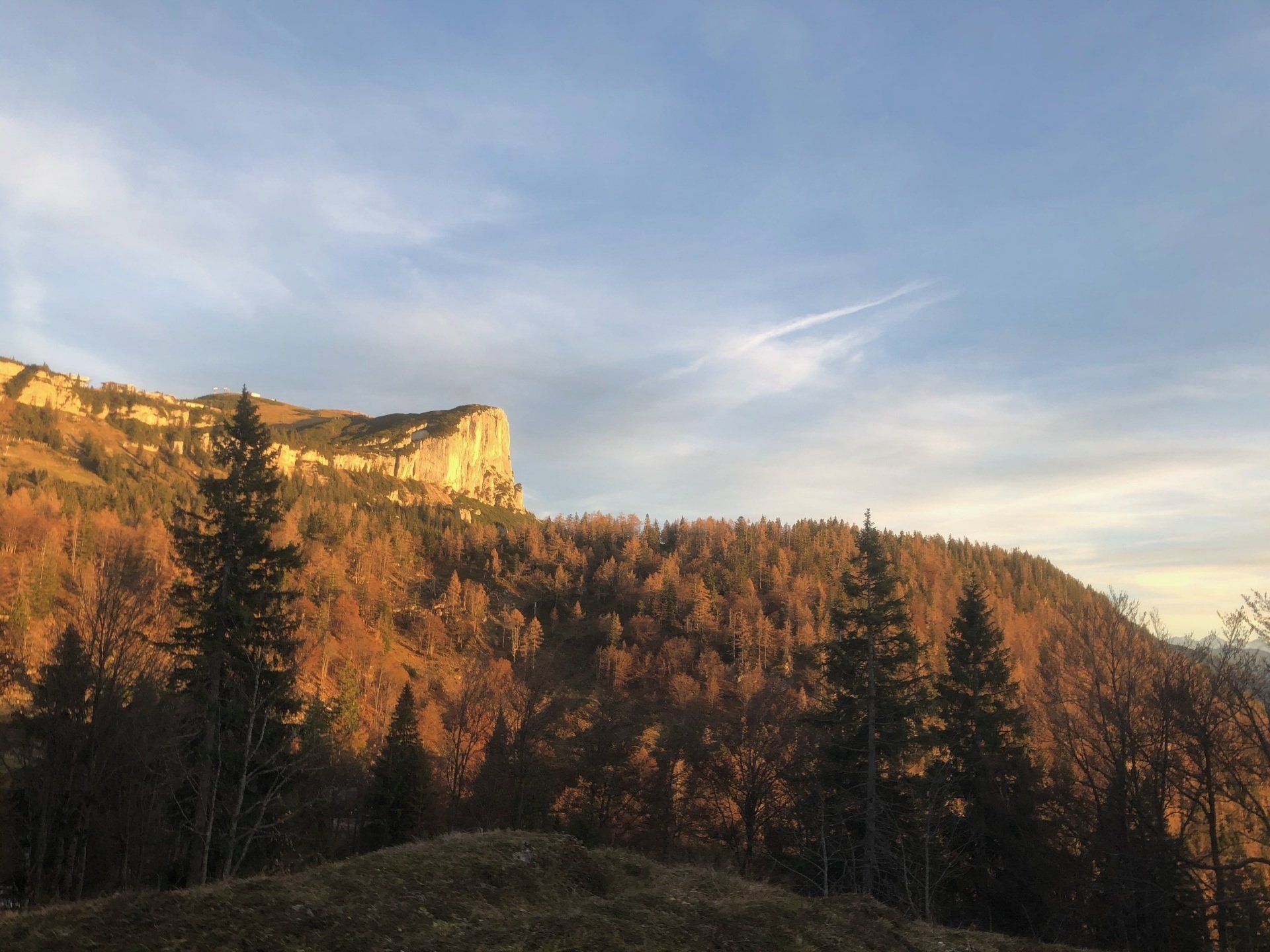Der Sonnenuntergang taucht eine Bergkette in goldenes Licht; Bäume im Vordergrund; blauer Himmel mit Kondensstreifen.