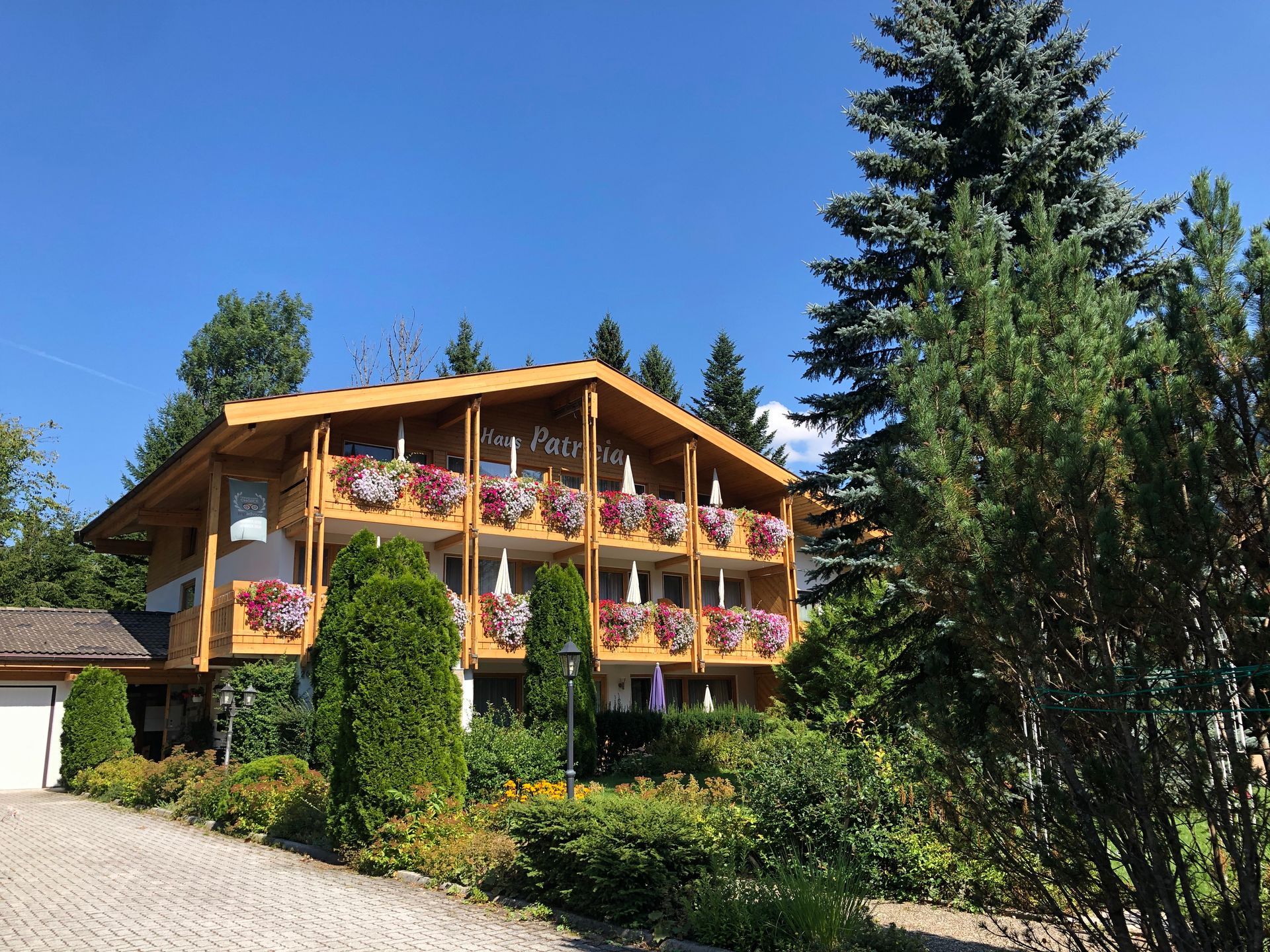 Dreistöckiges Holzhotel mit blumengeschmückten Balkonen, üppigem Grün und einer gepflasterten Auffahrt unter blauem Himmel.