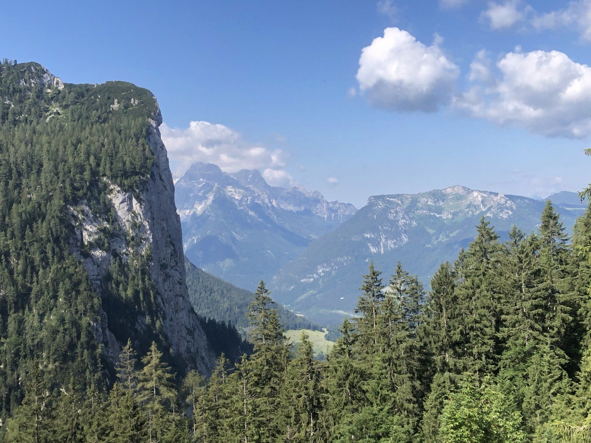 Berglandschaft mit Wald im Vordergrund, blauem Himmel mit Wolken und fernen Gipfeln.