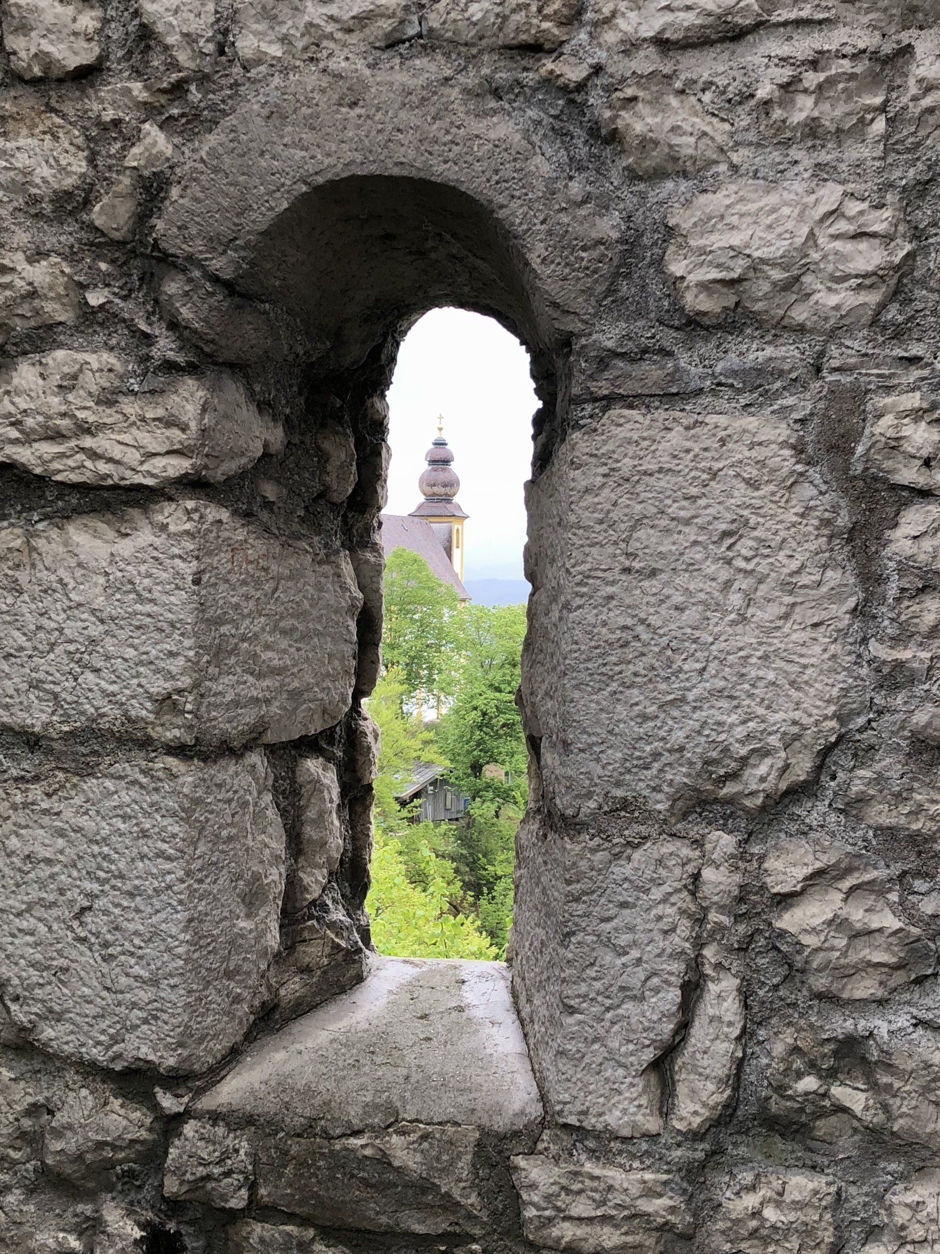 Kufstein: Steinmauer mit einer schmalen, bogenförmigen Öffnung, die ein entferntes Gebäude mit Turm einrahmt; grünes Laubwerk ist sichtbar.