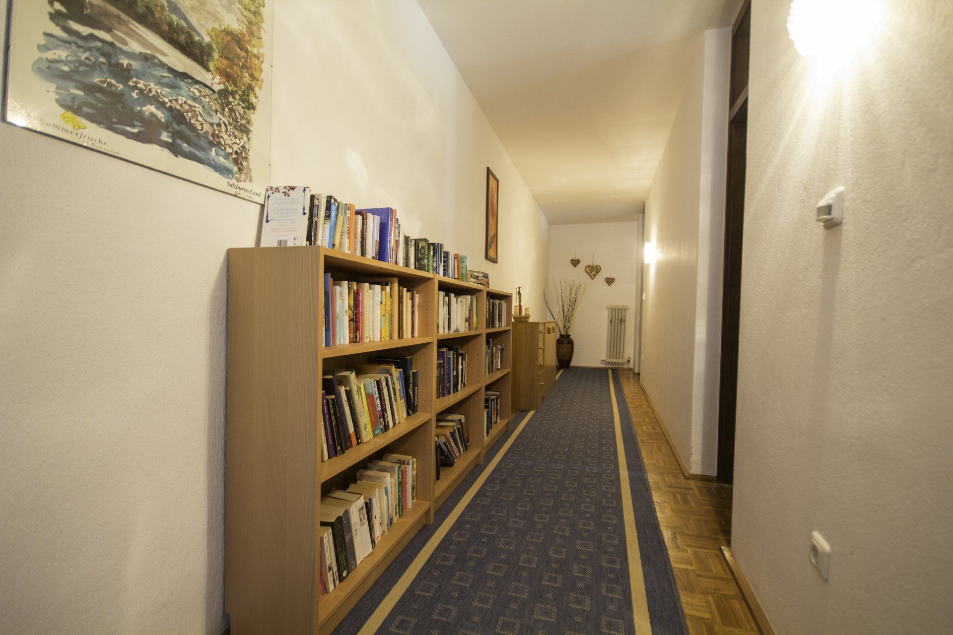 Schmaler Flur mit Bücherregal, Büchern und Kunstwerken. Blauer Teppichläufer.