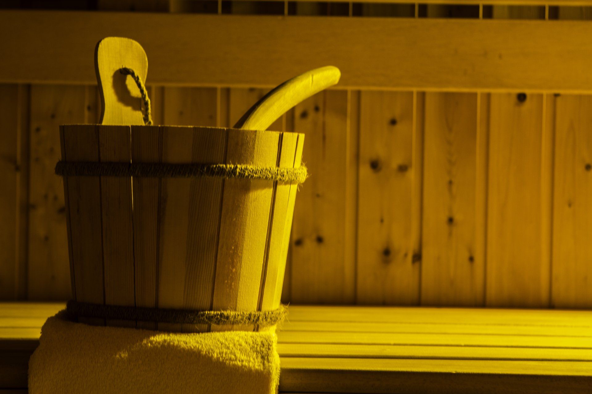 Saunaeimer aus Holz mit Schöpfkelle darin, auf einem Handtuch, in einer holzgetäfelten Saunalandschaft.