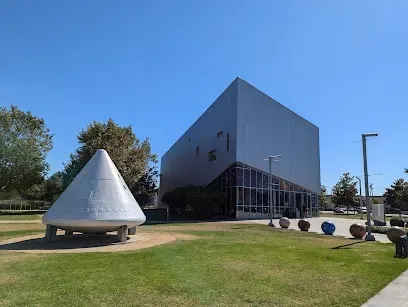 Tree Service Elmonte-Outdoor scene with a conical metallic sculpture on a grassy area in front of a modern, angular building. The sky is clear and sunny, creating a serene atmosphere.
