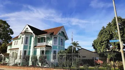 Tree Service El Monte Ca - A Victorian-style, mint green house with white trim stands under a blue sky. Surrounded by trees and a metal fence, it exudes a serene suburban charm.