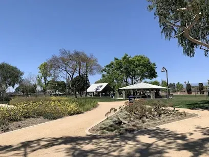 Tree Service Elmonte-A sunny park scene with a winding path, flowers, and trees. Two picnic shelters are visible in the background under a clear blue sky. Peaceful atmosphere.