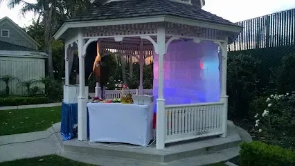 Tree Service Elmonte-A decorated white gazebo with a table, set in a garden. Soft colored lights inside create a festive atmosphere. Pathways and greenery surround it.