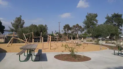 Tree Service Elmonte-Playground with wooden climbing structures on sand, surrounded by trees. Picnic tables on concrete path under a clear blue sky, creating a serene atmosphere.