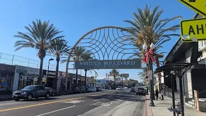 Tree Service El Monte Ca - Wide street view of Whittier Boulevard, featuring a decorative arch, palm trees, and small shops. The scene is bright and sunny, conveying a lively atmosphere.