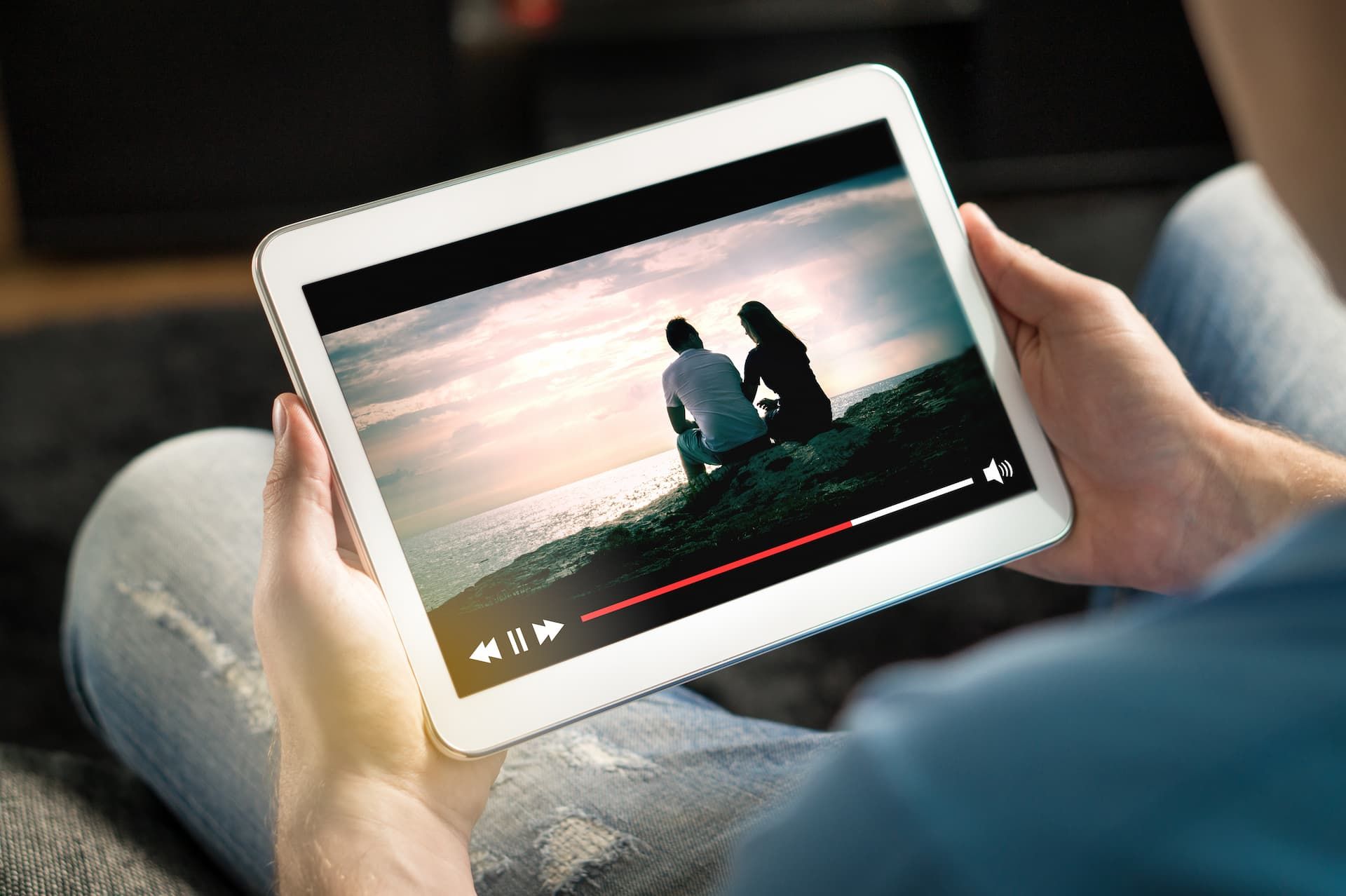 Hands holding a tablet playing a video of two people sitting by water at sunset