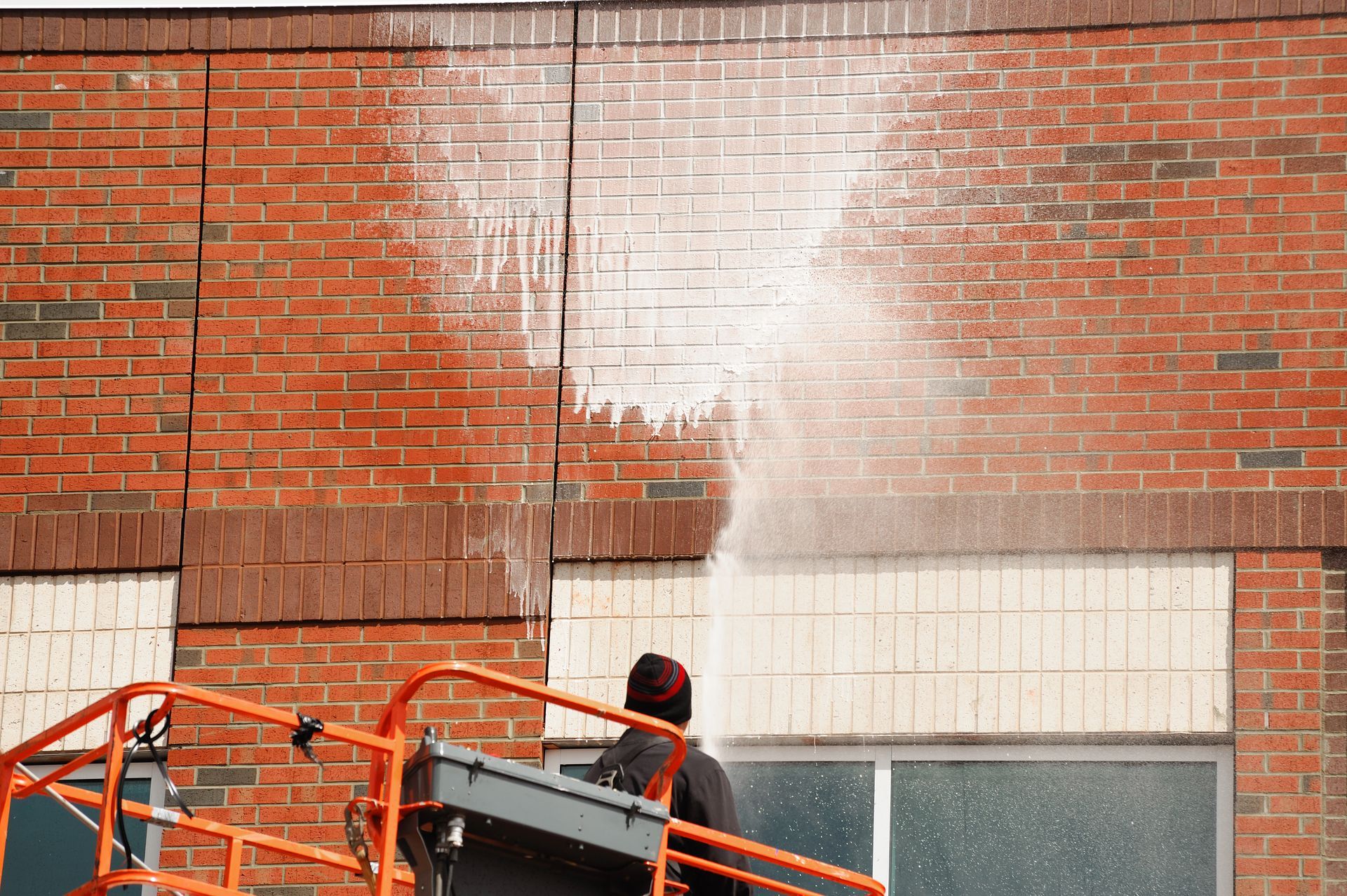 A worker on a lift power-washes a brick building facade with a high-pressure water spray.