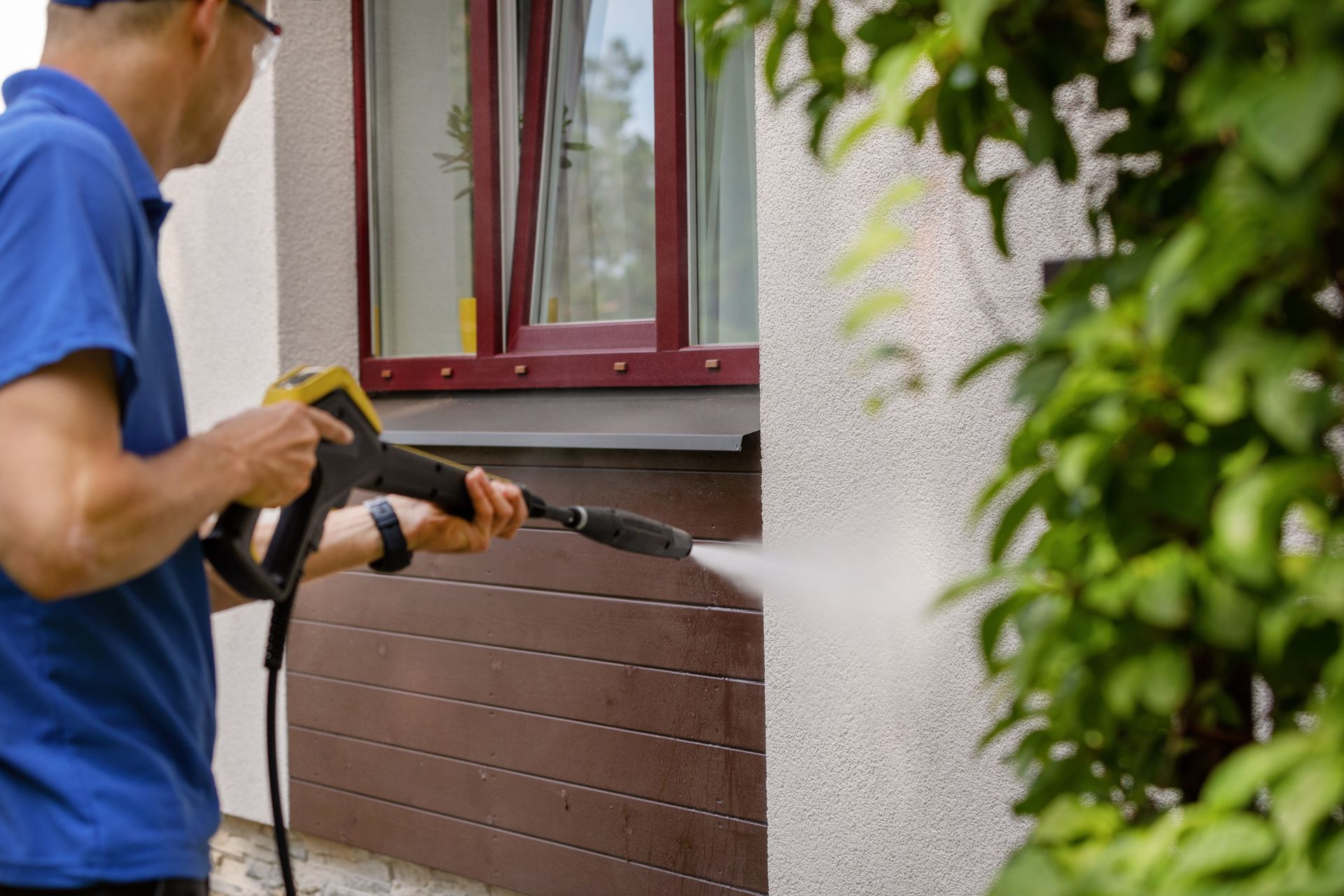 A person wearing a blue shirt uses a pressure washer to clean the exterior wall of a building near a window.
