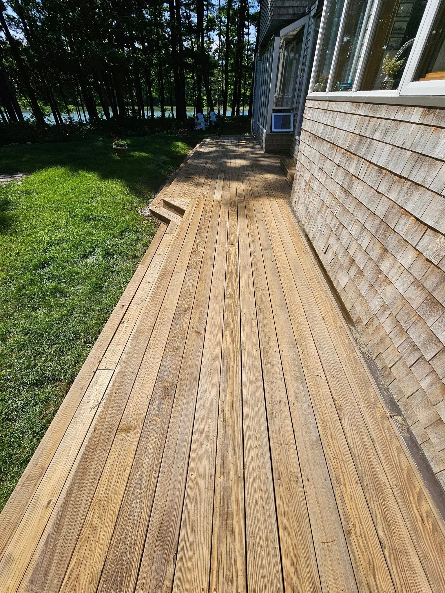A person in a plaid shirt uses a roller to stain wooden deck planks dark brown outdoors on a sunny day.