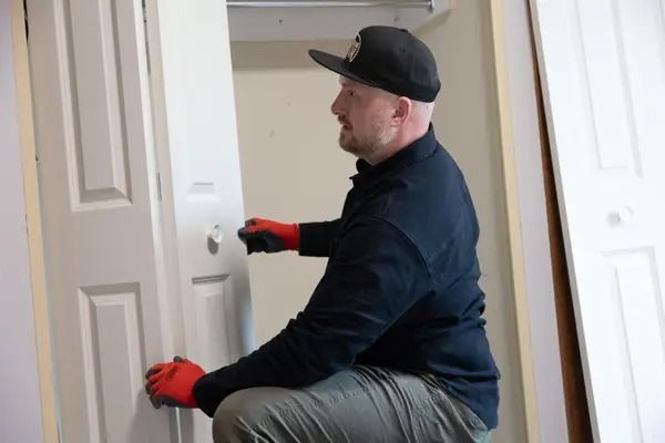 Man in black cap and gloves kneeling to install a door knob on a white door
