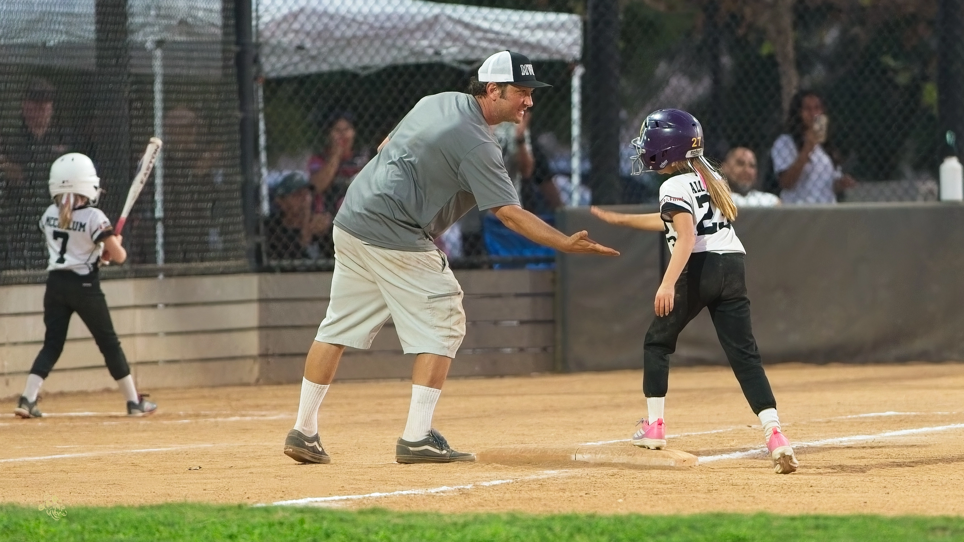 Coach high-fives a softball player on the field; another player bats.