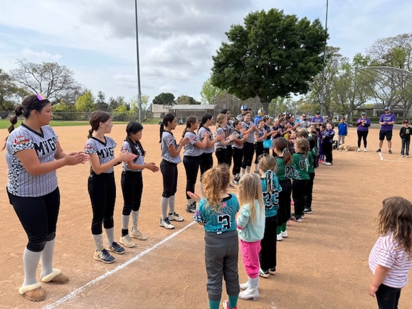 Softball teams lining up to clap before a game on a dirt field. Girls in team uniforms.