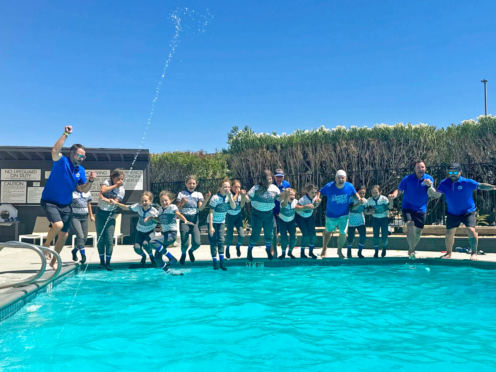 Group of people in blue shirts jumping into a pool on a sunny day. Water splashing.