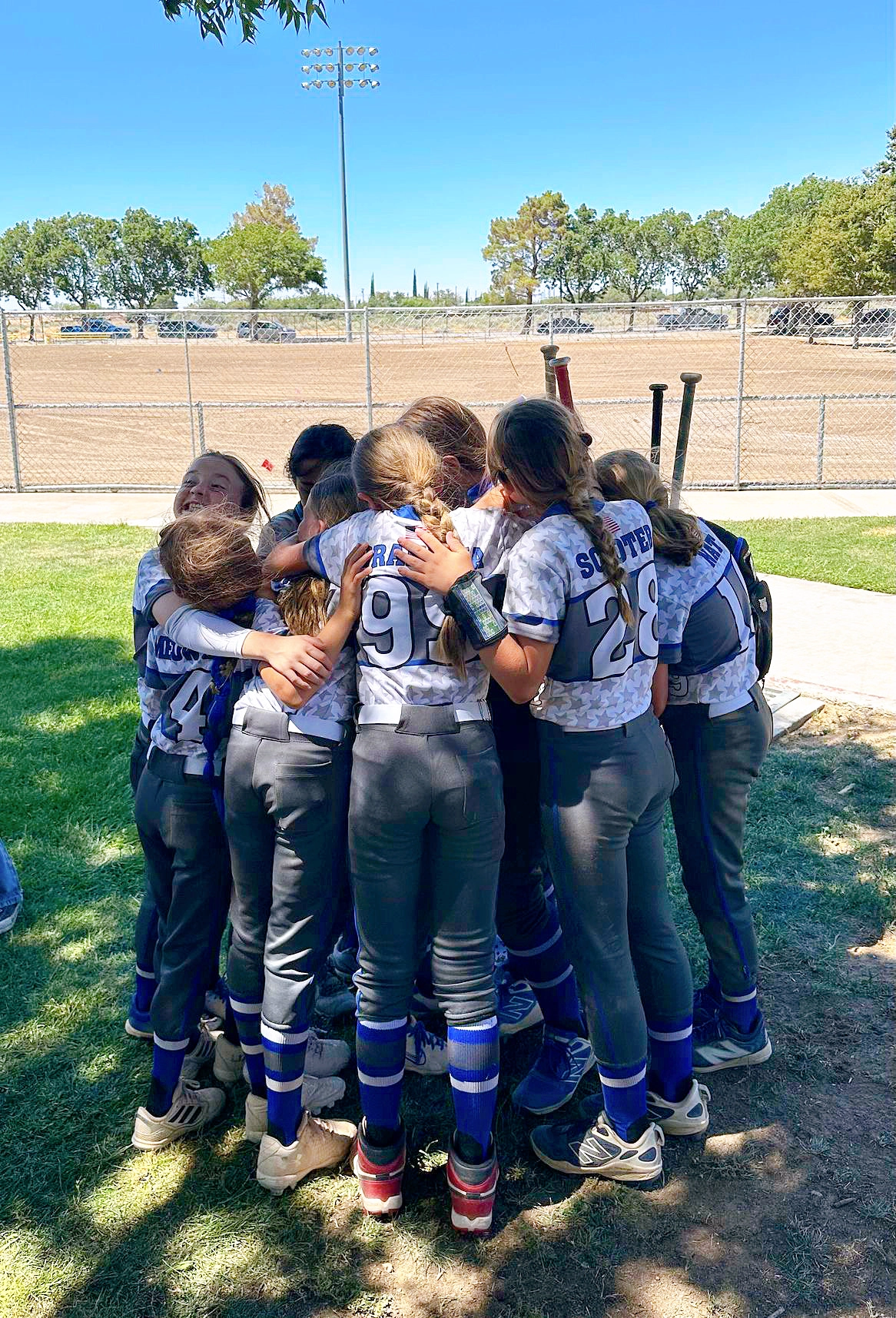 Softball team in gray and blue uniforms huddle together on a sunny field.