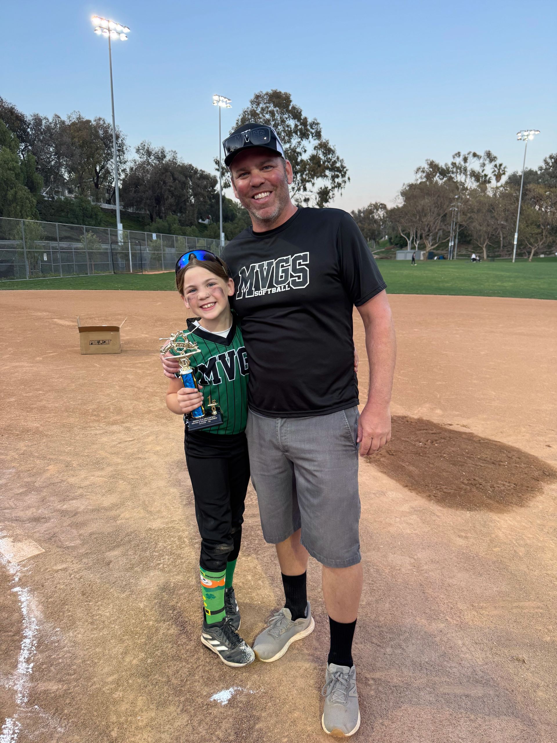 Man and child on a baseball field, smiling. The child holds a trophy. Green and black team uniform. Outdoors.