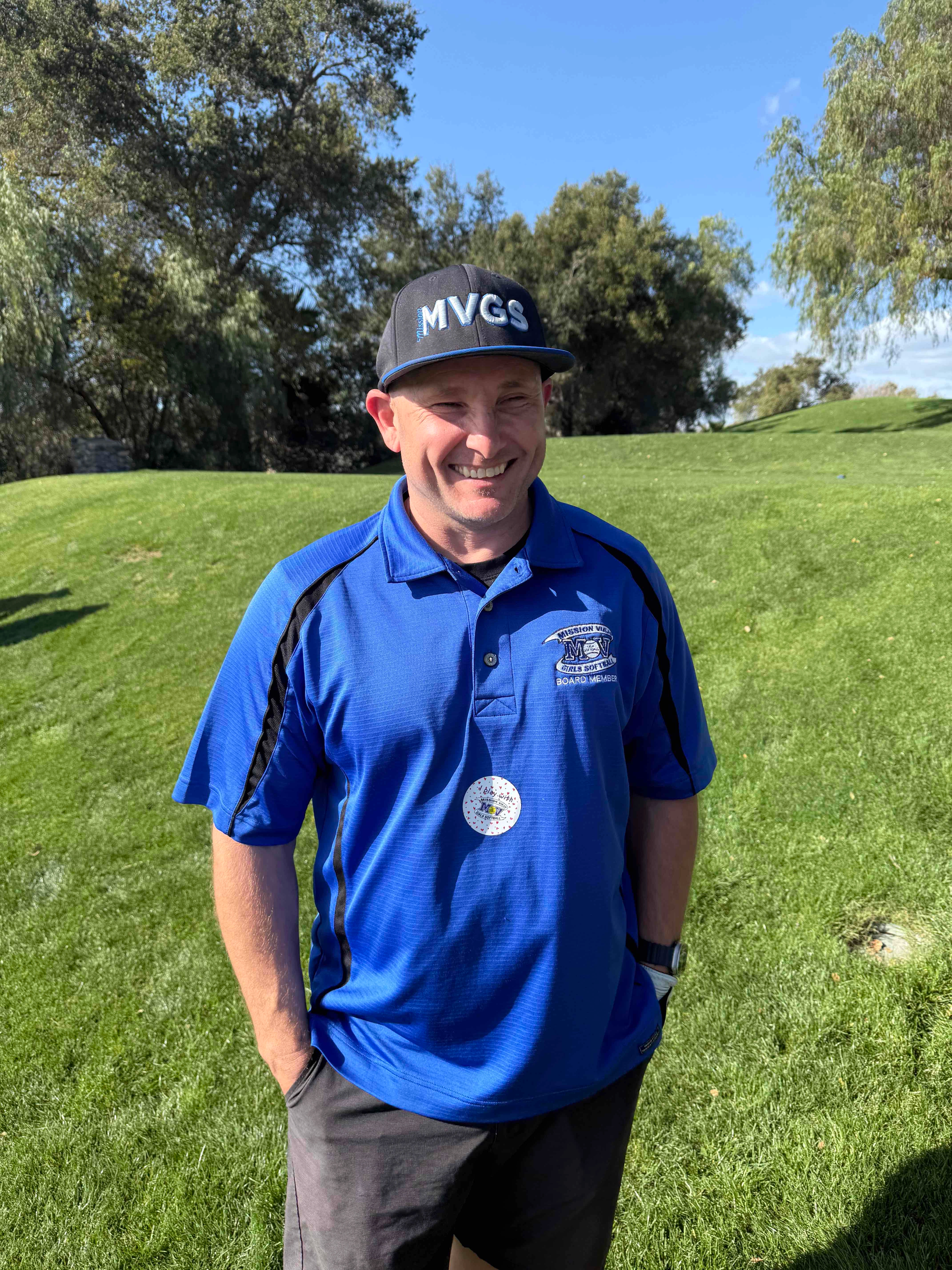 Man in blue shirt and black cap stands on a grassy field, smiling.