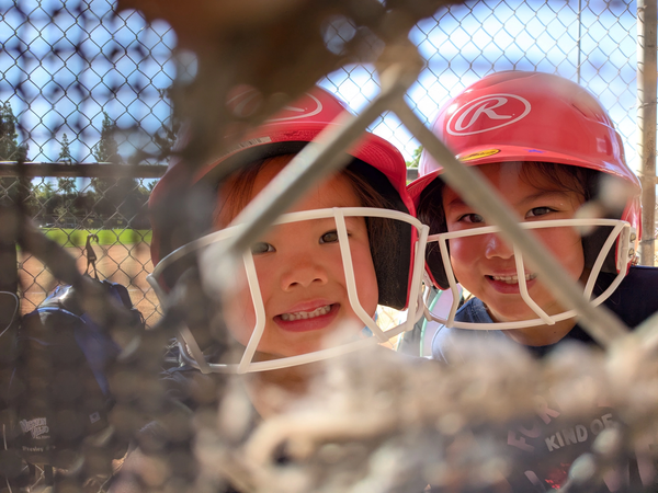 Two children in red baseball helmets with faceguards, peering through a chain-link fence.