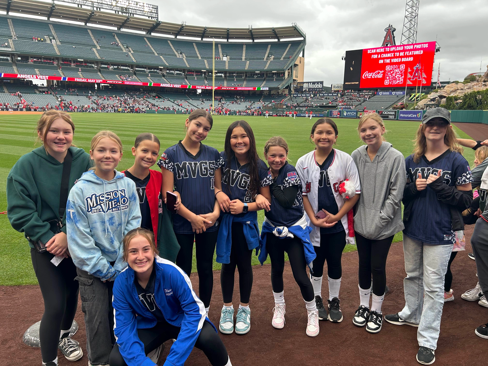 Group of girls pose on a baseball field. Fans in the stands, stadium in background.