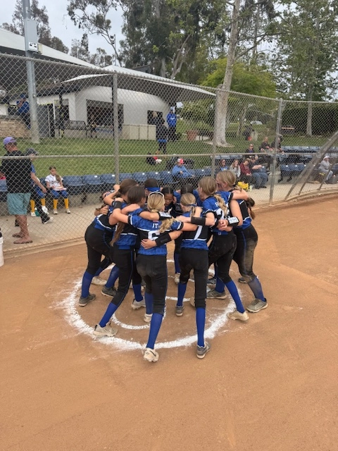 Softball team huddling on the infield, wearing blue and black uniforms, with a fence and building in the background.